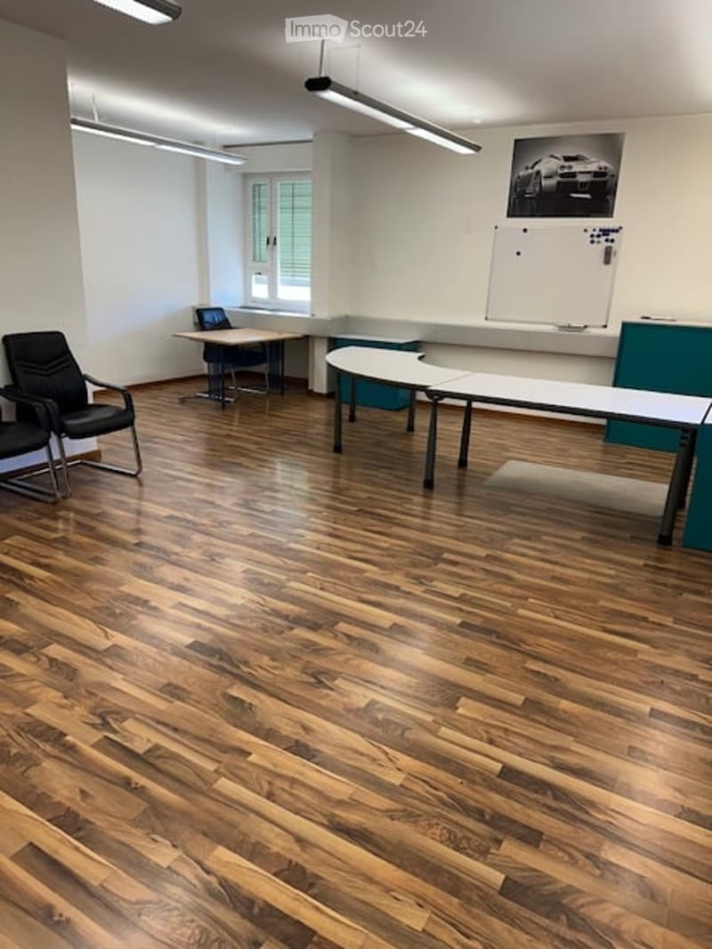 Bright room with wooden flooring, white walls, and large windows. Contains several black chairs, tables, a laptop, and a whiteboard.