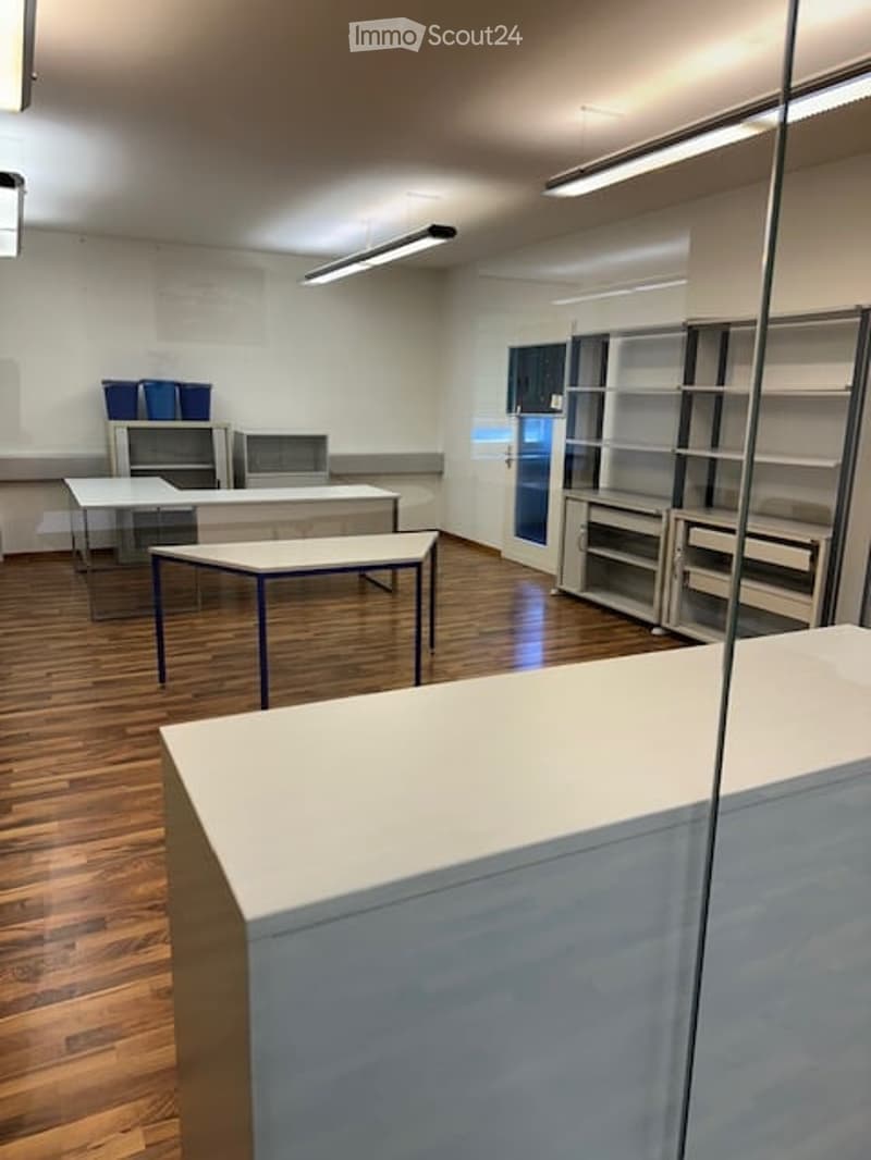 Bright, modern room with wooden flooring, white walls, and several tables and chairs arranged for a workspace or meeting area. Shelving units and cabinets are present along one wall.