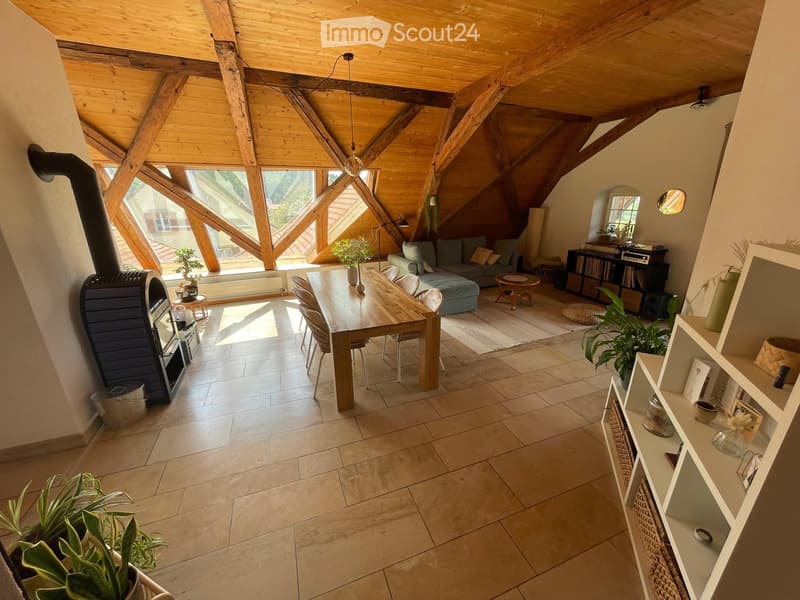 Open-plan living and dining area with exposed wooden beams, large windows, a sofa, dining table, chairs, bookshelf, and a fireplace.