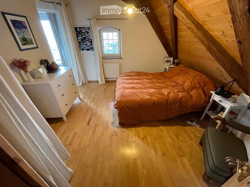 Cozy attic bedroom with wooden floors, slanted wooden ceiling beams, and large windows. Features a bed with orange bedding, a nightstand, a dresser, and a chair.