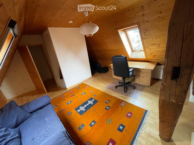 Cozy attic room with wooden walls and floors, featuring a blue sofa, a wooden table with a colorful rug, a desk with a chair, and a hanging paper lantern light fixture.