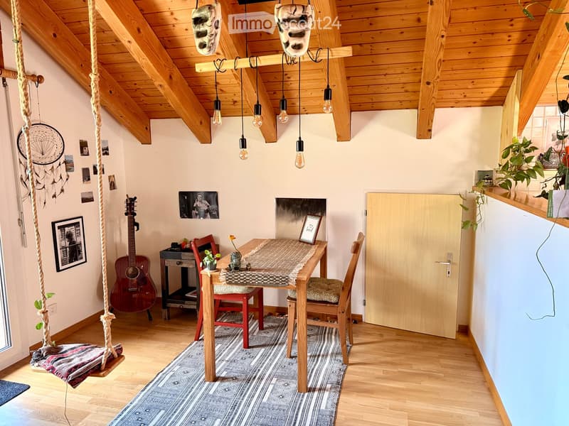 Cozy attic room with wooden beams, exposed ceiling, hanging light bulbs, a small table with chairs, a guitar, a dreamcatcher, and a swing seat.