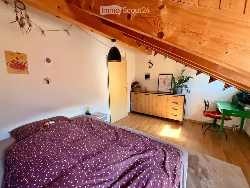 Cozy attic bedroom with wooden slanted ceilings, a bed with purple bedding, a small desk with a chair, a wooden cabinet, and decorative items on the walls.