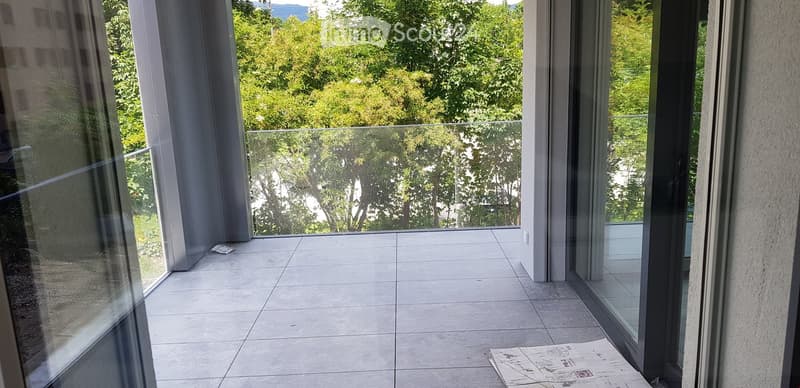 Modern balcony with large glass railings, tiled floor, and a scenic view of green trees and hills.