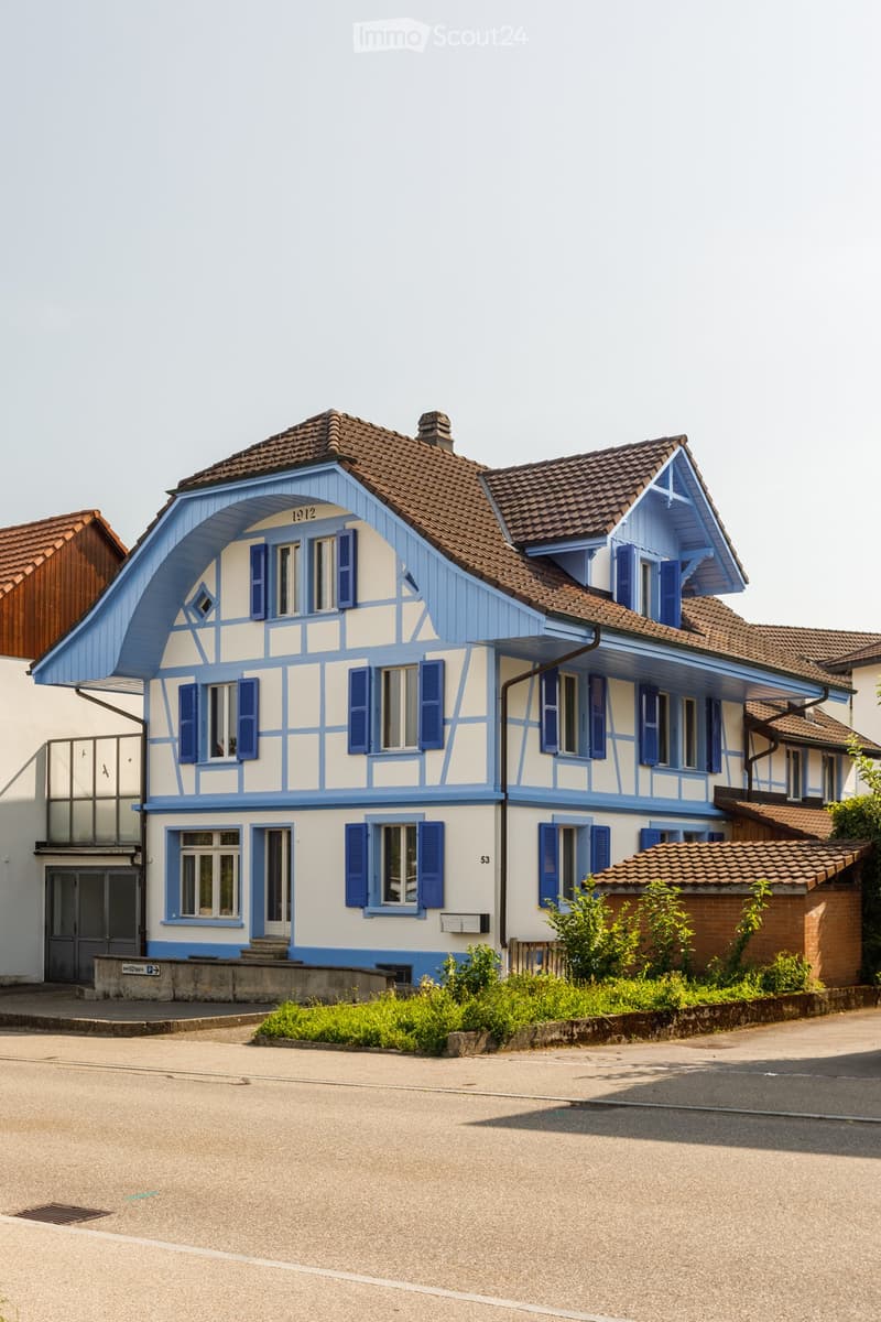 Two-story house with blue and white exterior, brown tiled roof, multiple windows with shutters, and small garden area.