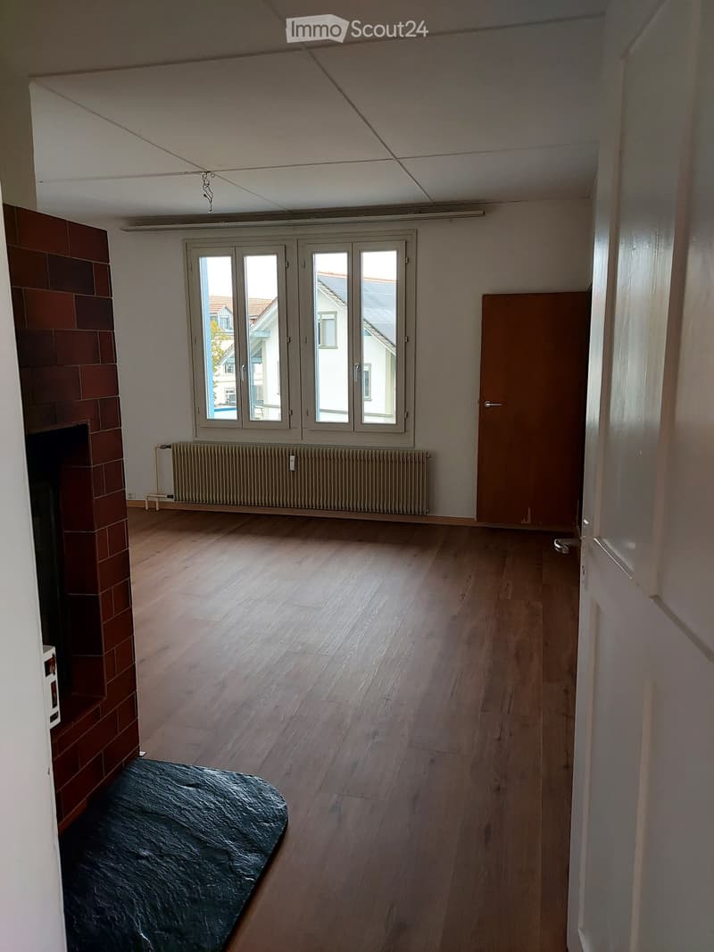 Empty room with wooden flooring, large windows, radiator, and a fireplace with brick tiles.