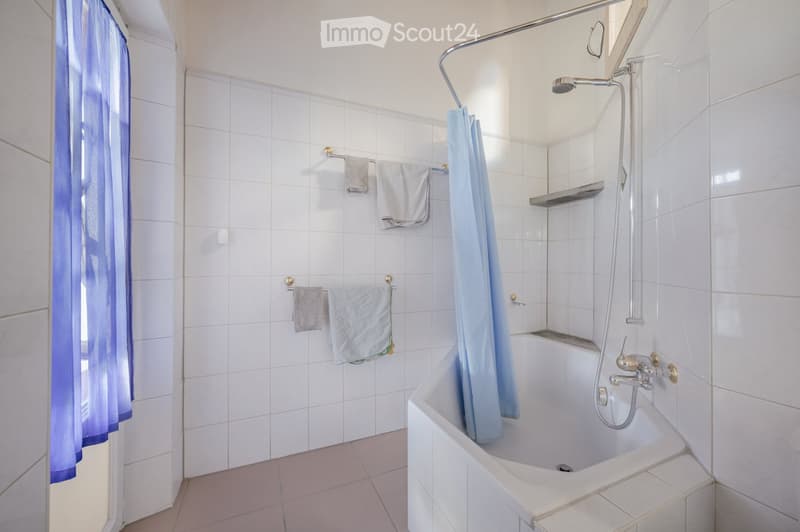 A clean, modern bathroom with white tiled walls and floor, a bathtub with a shower curtain, towel racks with folded towels, and a showerhead with a handheld sprayer.
