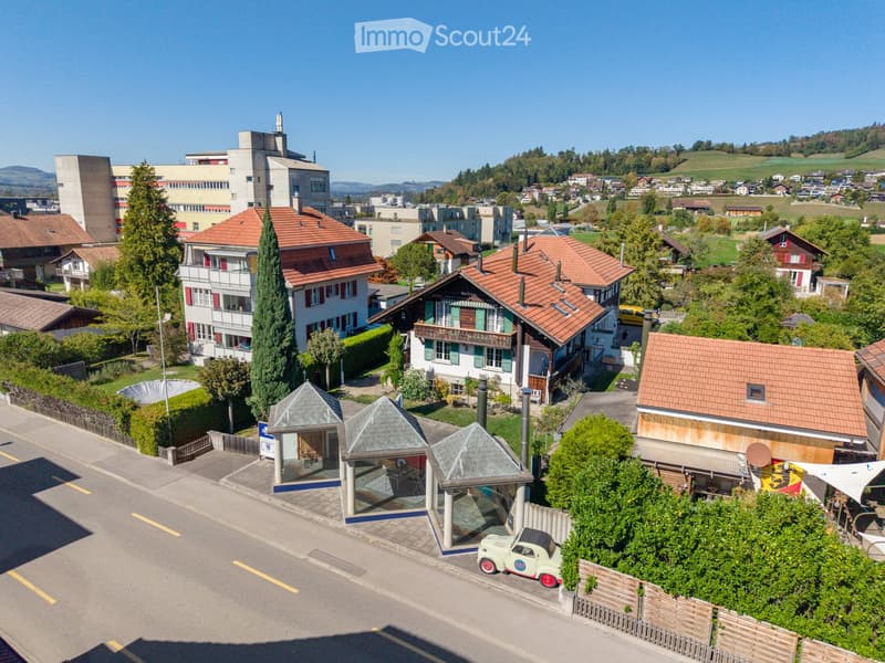 Aerial view of a residential area with multiple houses, red-tiled roofs, and surrounding greenery. Includes a small gazebo structure and a vintage car parked nearby.