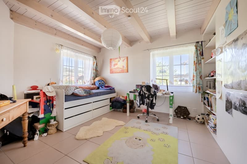 Cozy children's bedroom with a lofted bed, desk, shelves, and colorful rugs. The room features wooden beams on the ceiling and large windows with curtains.