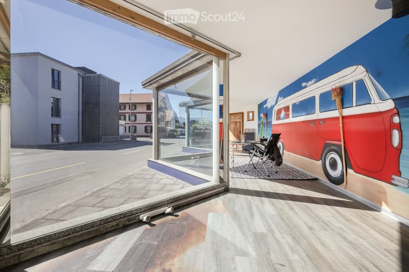 Bright, modern room with large glass doors leading to an outdoor area, wooden flooring, and a mural of a van on the wall. Includes a small table, chairs, and a floor lamp.