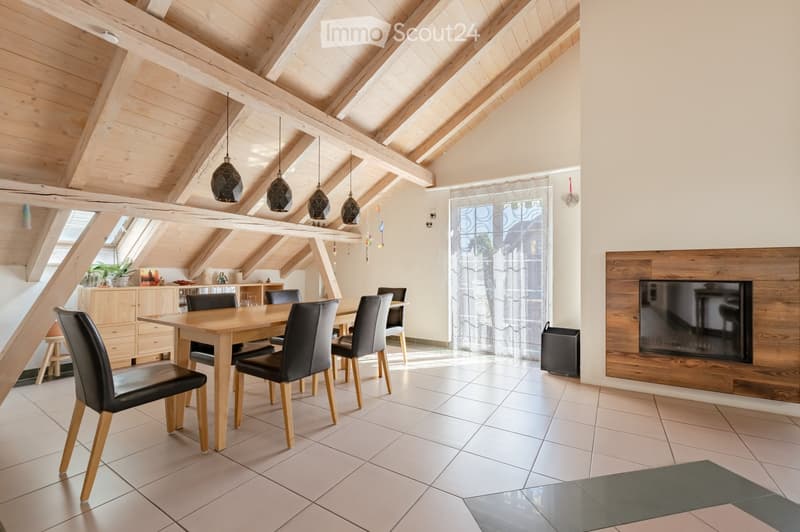 Bright, open dining area with exposed wooden beams, a large dining table surrounded by black leather chairs, a modern fireplace with a TV screen, pendant lights, and a small cabinet with decorative items.