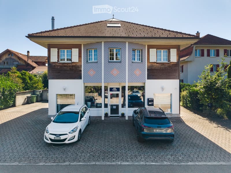 Two-story building with a tiled roof, white and blue facade, and a storefront with glass windows and a sign. Two cars are parked in front of the building on a paved driveway.