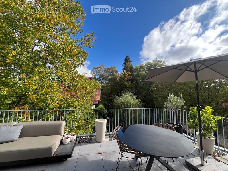 Balcony with a sofa, table, chairs, and an umbrella, surrounded by potted plants and trees.