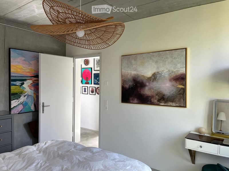 Modern bedroom with a large bed, a vanity table with a mirror, a hanging woven ceiling light, and several artworks on the walls.