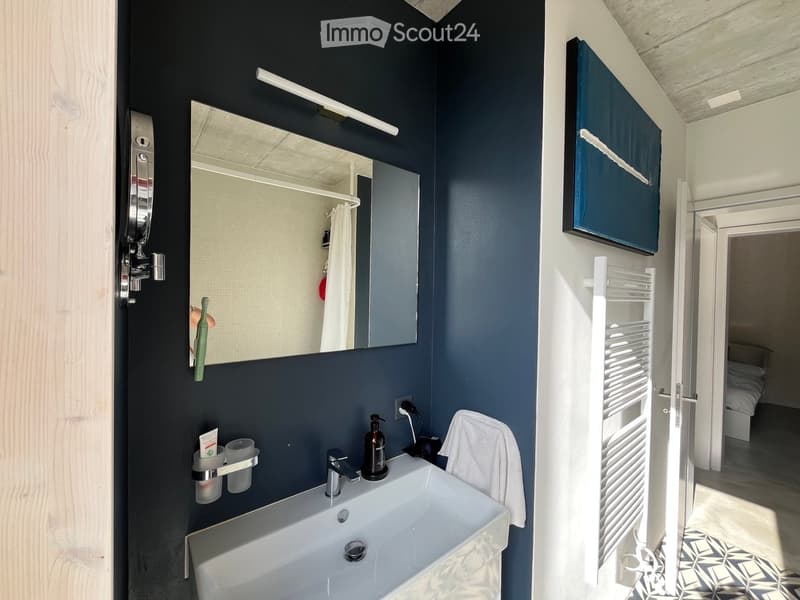 Modern bathroom with dark blue walls, white sink, mirror, toothbrush, towel, radiator, and a view into an adjacent bedroom.