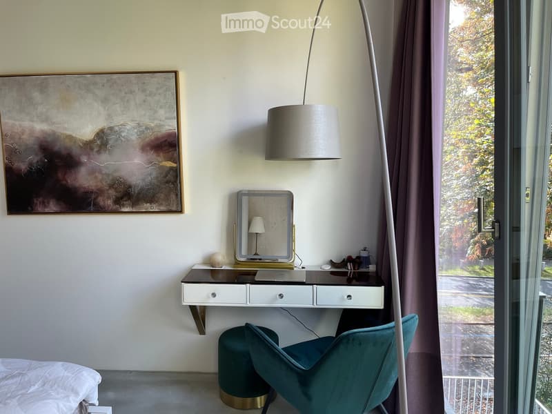 Modern bedroom with a bed, teal chair, round stool, desk with mirror, and a large abstract painting on the wall.