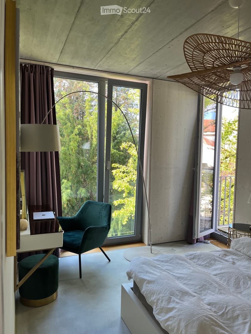 Modern bedroom with a large bed, teal armchair, round stool, desk with laptop, floor lamp, and large windows with a view of greenery.