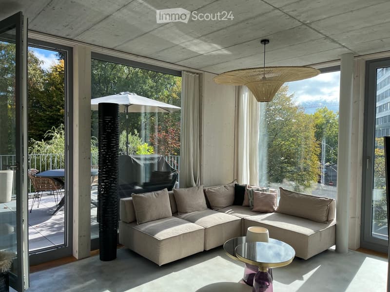 Modern living room with large windows, a sectional sofa, coffee table, floor lamp, and outdoor furniture visible through the glass doors.