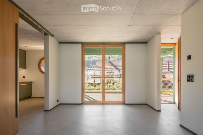 Empty room with white walls, concrete floor, and large sliding glass doors leading to an outdoor area. Minimalist design with a small cabinet and round mirror visible on the left side.
