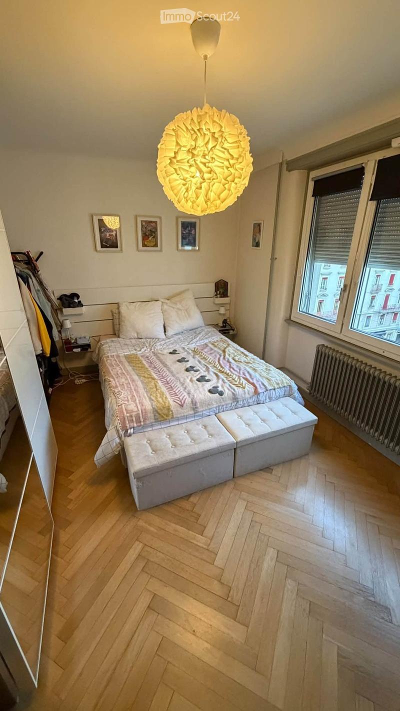 Cozy bedroom with wooden herringbone flooring, a large bed with patterned bedding, a matching bench at the foot, a modern hanging lamp, and several picture frames on the wall.