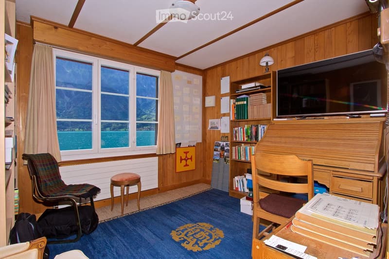A cozy, wood-paneled room with large windows overlooking a scenic lake, featuring a comfortable chair, a small stool, a bookshelf filled with books, a television, and a wooden table with papers and a remote control.