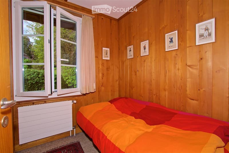 Cozy bedroom with wooden walls, a large window with white frames and curtains, a bed with a colorful orange and red duvet, a radiator below the window, and several framed pictures on the wall.