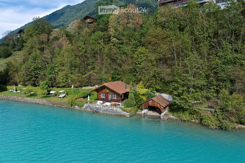 A serene lakeside scene with a small wooden house, a dock, lounge chairs, and a boat house, surrounded by lush greenery and mountains.