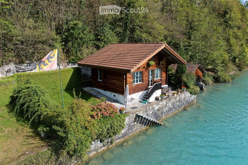 A wooden house with a tiled roof, situated near a river with a small stone platform and outdoor seating area.