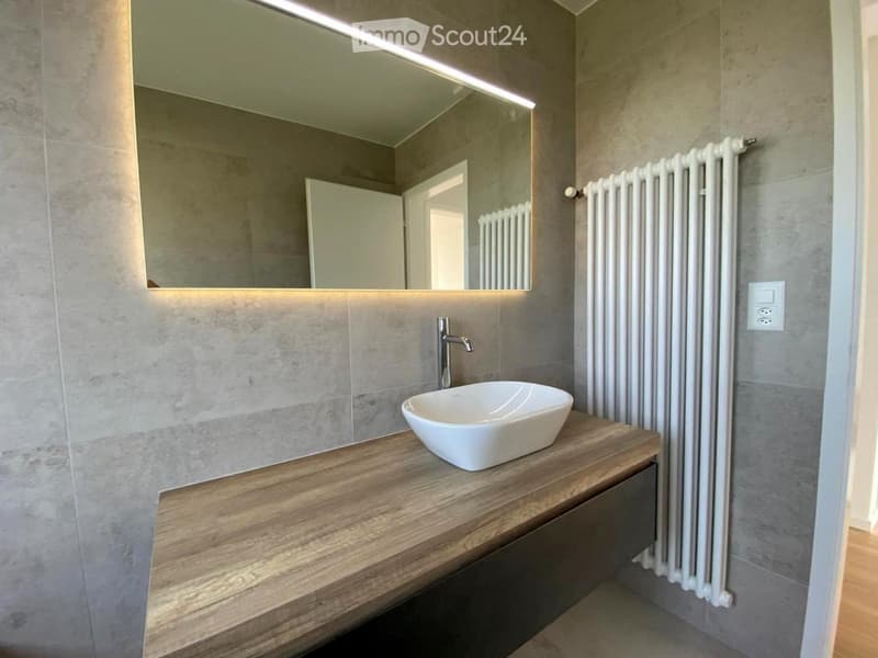 Modern bathroom with a wooden vanity, white sink, sleek faucet, large mirror with integrated lighting, and a white radiator.