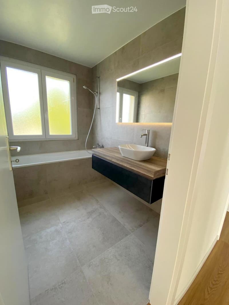 Modern bathroom with a sleek wooden vanity, white sink, bathtub, and large mirror with integrated lighting. The space features light grey tiles and a contemporary design.
