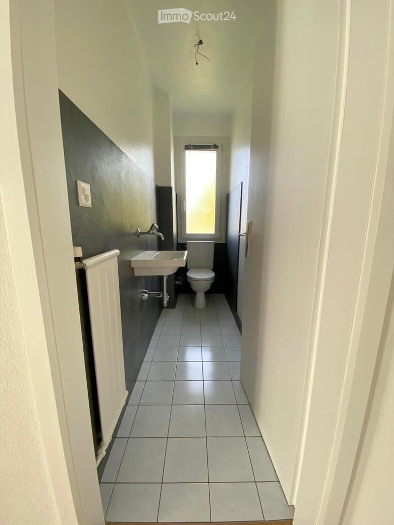 Small bathroom with white tiled floor, white walls, a sink, and a toilet. Features a window with frosted glass and a radiator on the left side.