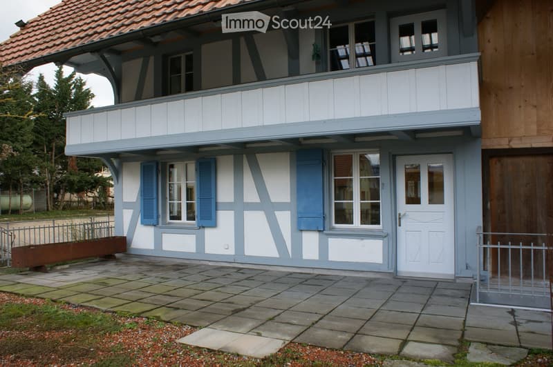 Two-story house with white and grey exterior, blue shutters, and a balcony on the upper floor. The entrance features a white door with glass panels, and the ground is paved with stone tiles.