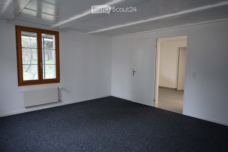 Empty room with white walls, grey carpet, a window with wooden frame, radiator, and an open doorway leading to another room.