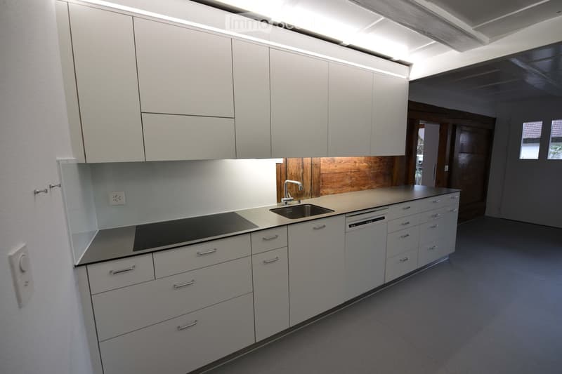 Modern kitchen with white cabinets, stainless steel sink, dishwasher, and electric stove. Wooden backsplash and grey floor.