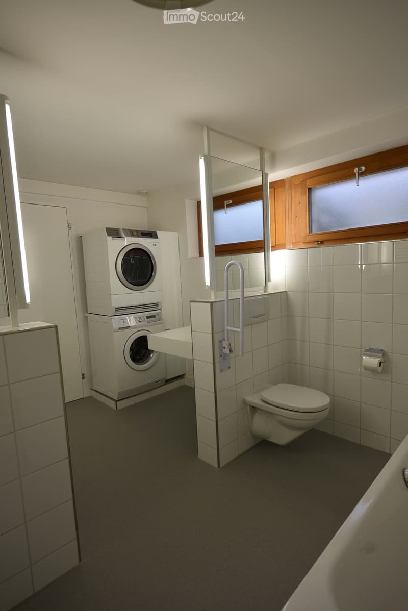 Modern bathroom with white tiled walls, a washing machine and dryer unit, a toilet, a bathtub, and a large mirror with built-in lighting.
