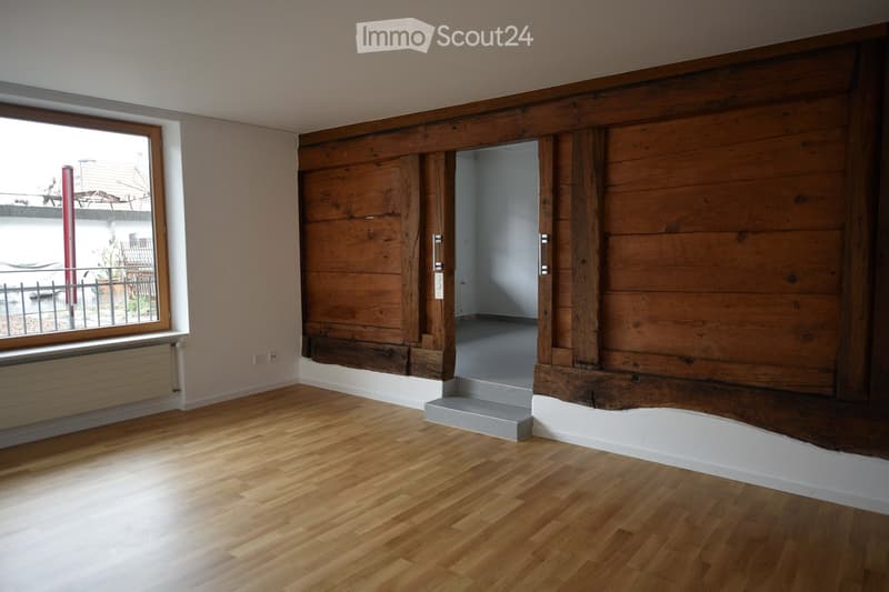 Empty room with wooden flooring, white walls, and a large window with a radiator below it. An open doorway leads to another room with a similar wooden floor.