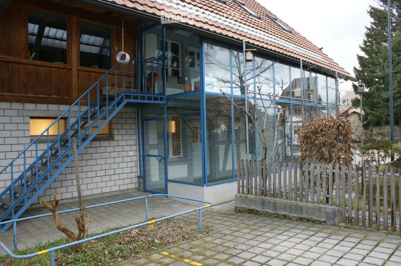 Two-story house with a red-tiled roof, a mix of wooden and brick exterior, large glass windows, and a blue metal staircase leading to an upper level.