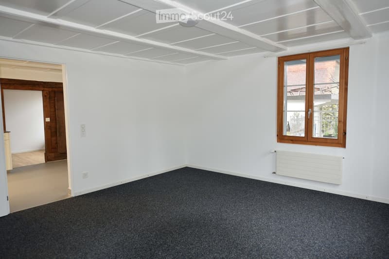 Empty room with white walls, grey carpet, a window with wooden frame, radiator, and a doorway leading to another room.