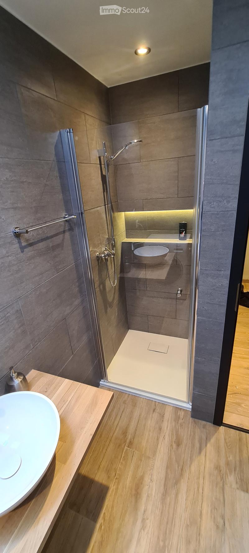 Modern bathroom with a wooden floor, a white sink on a wooden vanity, a glass-enclosed shower with a modern faucet, and a built-in shelf with a small bottle on it.