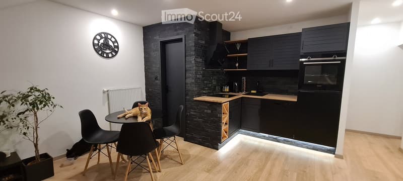 Modern kitchen with dark cabinets, wooden countertops, built-in oven, sink, and a small dining area with a table and chairs. A cat is resting on the table, and a potted plant is near the wall.