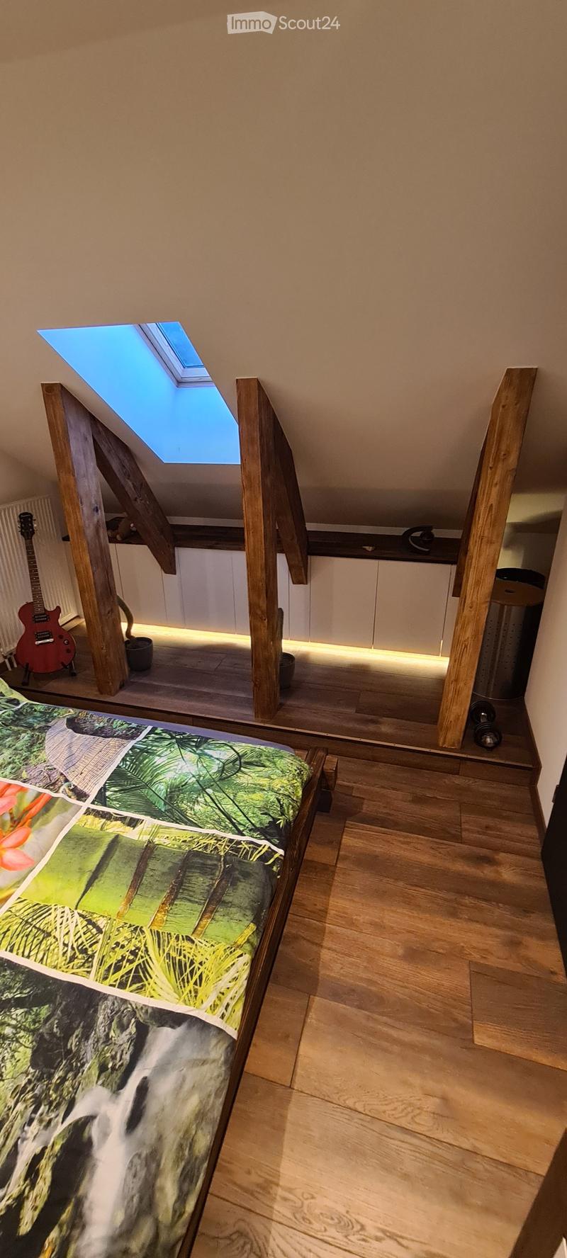 Bright attic bedroom with wooden floors, skylight window, bed with tropical-themed bedspread, and a red guitar leaning against the wall.