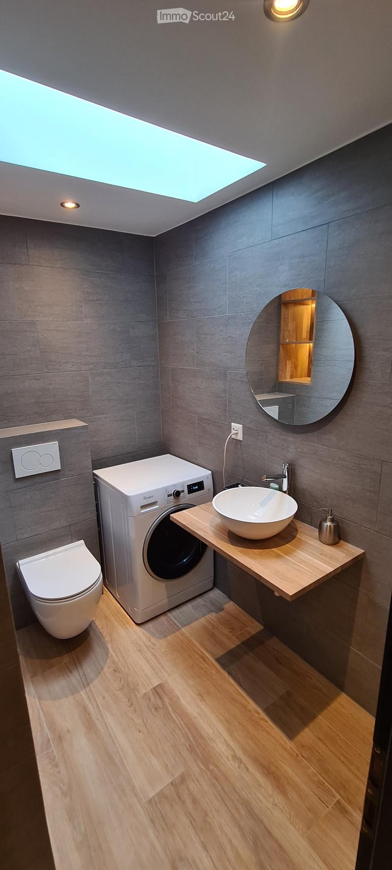 Modern bathroom with grey tiled walls, wooden floor, toilet, washing machine, sink, and a round mirror with built-in lighting.