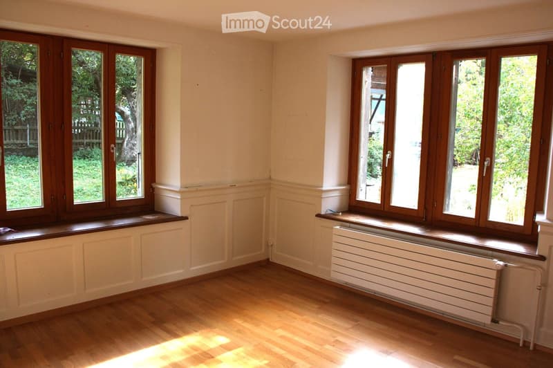 Empty room with wooden flooring, large windows with wooden frames, radiators beneath the windows, and white walls with decorative paneling.