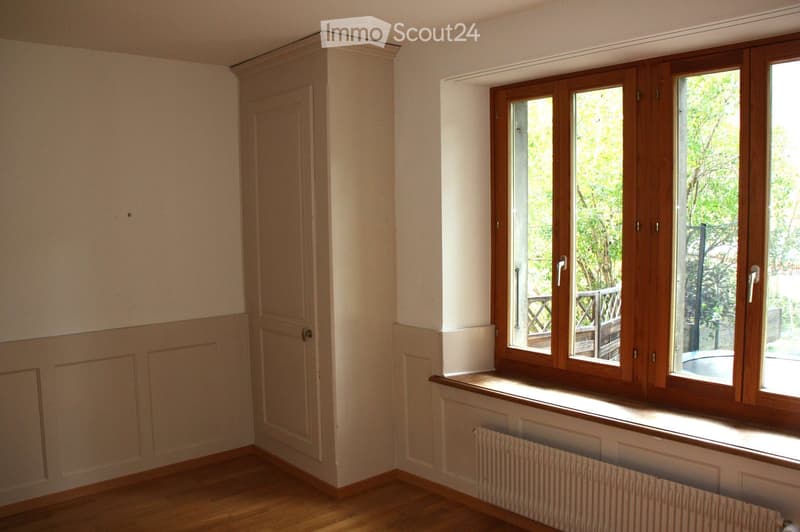Empty room with wooden flooring, white walls, a door, and large windows with wooden frames offering a view of greenery outside.
