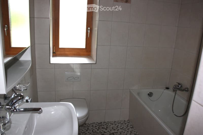 Bathroom with a white tiled wall, a sink with a chrome faucet, a toilet, and a bathtub with a handheld shower attachment. There are two windows with wooden frames and a small shelf above the sink.
