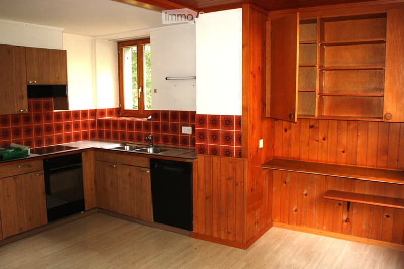The kitchen features wooden cabinets, a sink with a faucet, an oven, a dishwasher, and wooden shelves. The walls are paneled in wood, and the floor is light-colored wood.