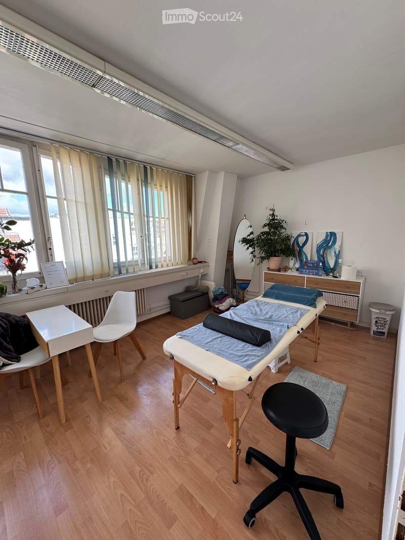 Well-lit room with wooden flooring, massage table, chair, small table, mirror, and potted plant near the window.