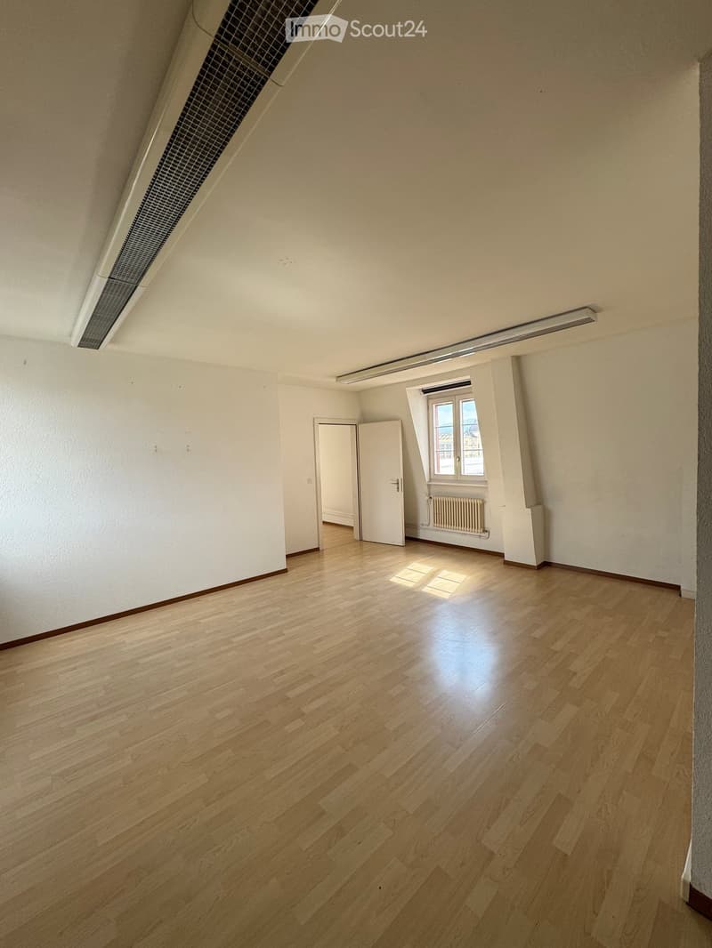 Empty room with wooden flooring, white walls, and a window with a radiator below it. The room has a door leading to another space and a ventilation duct in the ceiling.