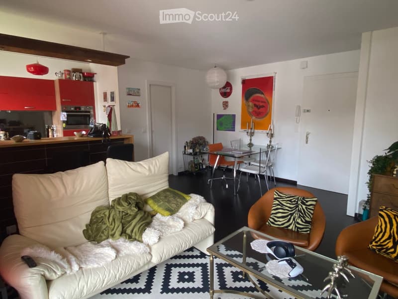 Modern living room with a white sofa, coffee table, dining area, and kitchen area. Features include a patterned rug, decorative pillows, and various furniture pieces.
