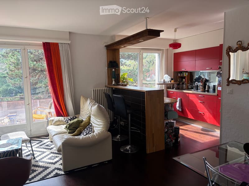 A modern, open-plan living and dining area with a cream sofa, glass coffee table, and red kitchen cabinets. The space features large windows with a view of greenery, a bar counter, and a dining table with chairs.